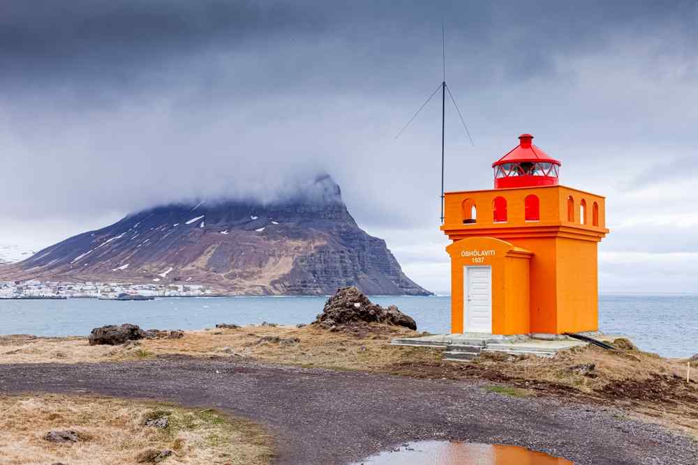 Bright orange lighthouse on the Westfjords coast with a misty mountain across the water.