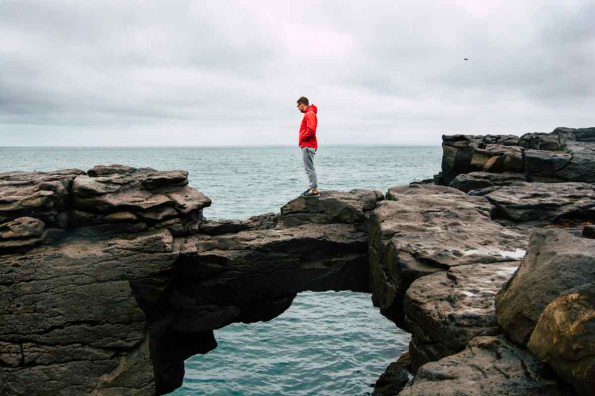 Solo traveler in a red jacket standing on a rocky natural bridge above the ocean, staring down at the crashing waves on a cloudy day