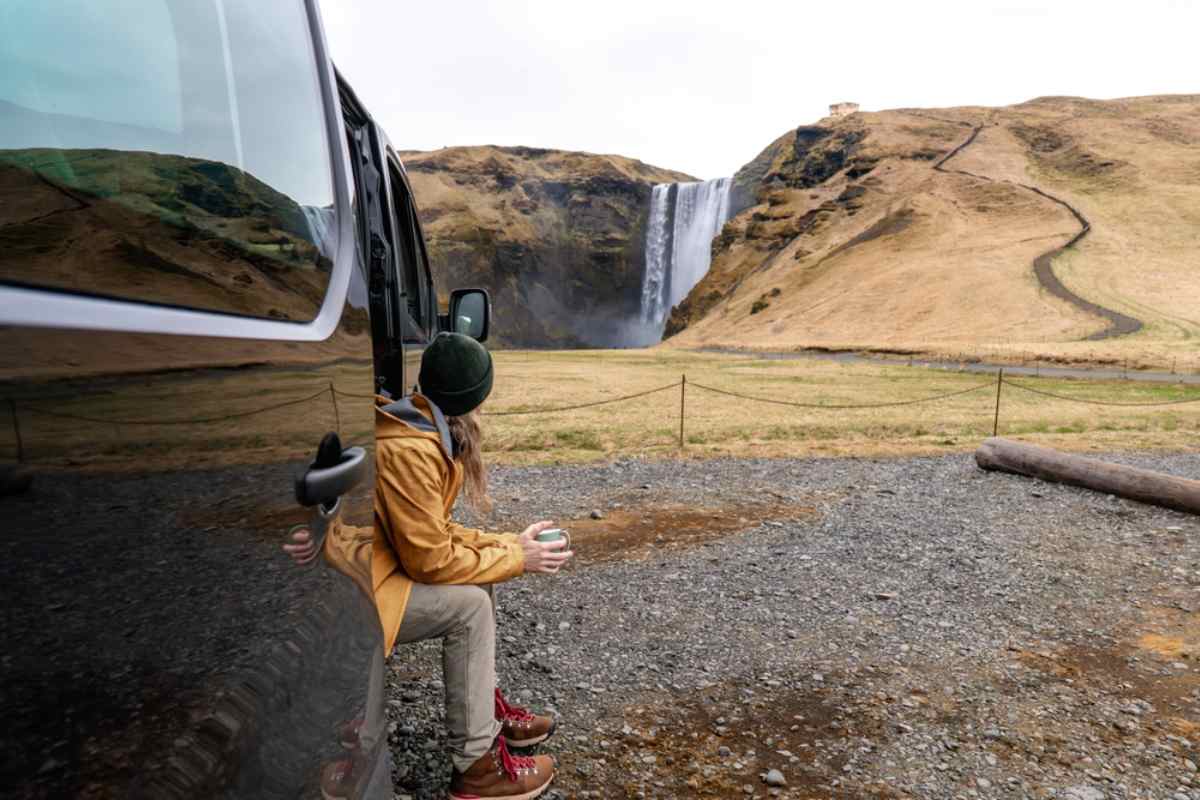 Camper sitting in the doorway of a parked van, drinking from a mug while looking at a tall waterfall cascading down golden cliffs in Iceland