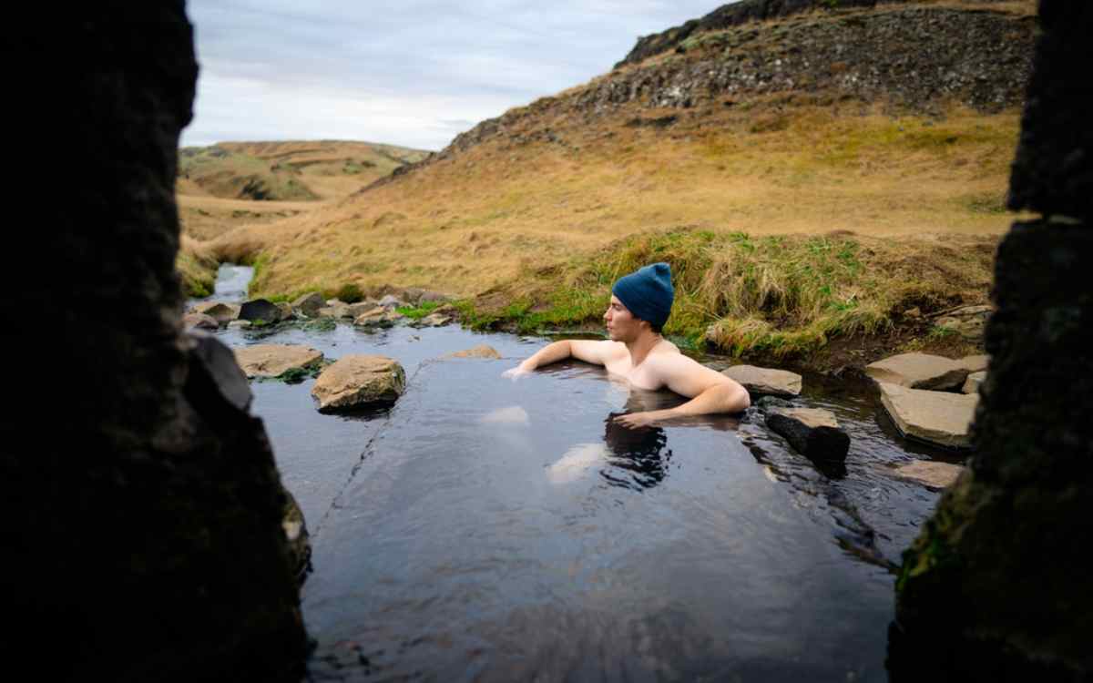 Traveler relaxing alone in a steaming natural hot pool in the countryside, wearing a blue beanie and looking out over mossy hills