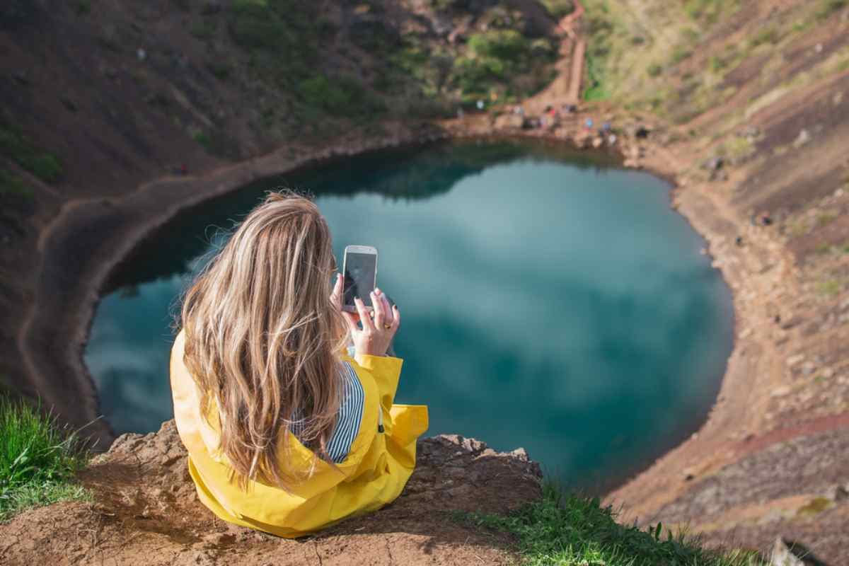 Young woman in a yellow raincoat sitting on the rim of a crater lake, taking a picture with her phone of the turquoise water below