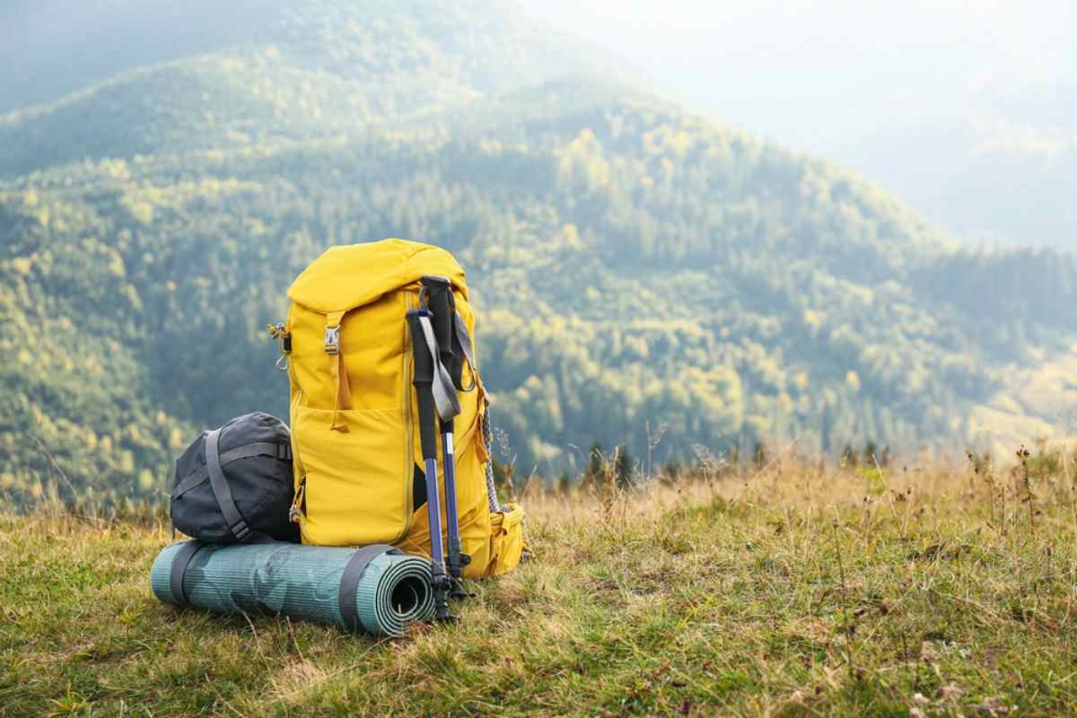 Yellow hiking backpack, trekking poles and rolled sleeping mat resting on a grassy hilltop above a green forested valley on a sunny summer day