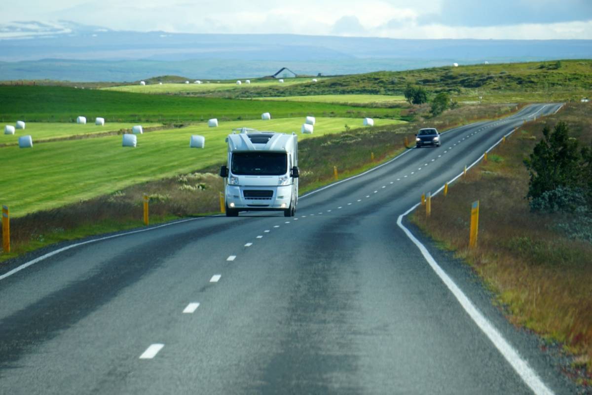 Motorhome driving Iceland’s Ring Road past green farmland and hay bales on a bright summer day.