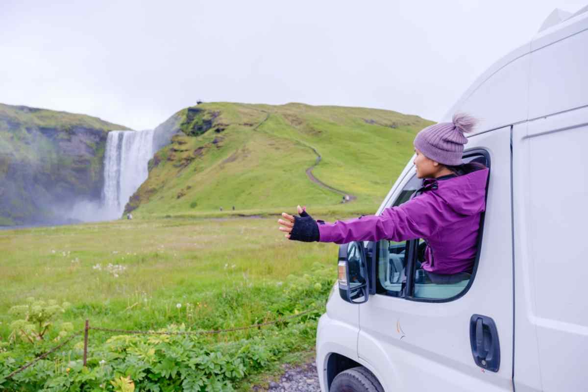 Traveler reaching out from a white campervan toward a tall waterfall across a green Icelandic valley.